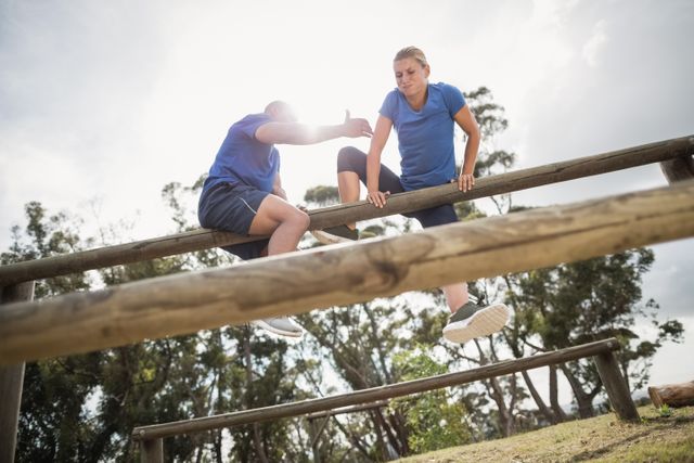 Man helping woman climb over a wooden hurdle during an outdoor boot camp. Ideal for illustrating teamwork, fitness training, and motivational themes. Suitable for use in articles, advertisements, and promotional materials related to physical fitness, outdoor activities, and team-building exercises.