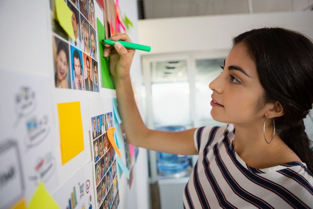 Young businesswoman writing on sticky note at creative office, ideal for depicting teamwork, brainstorming sessions, and creative planning. Useful for business, innovation, and project management themes.