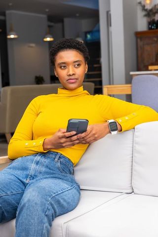 African american woman sitting on sofa holding phone wearing mustard top and smartwatch