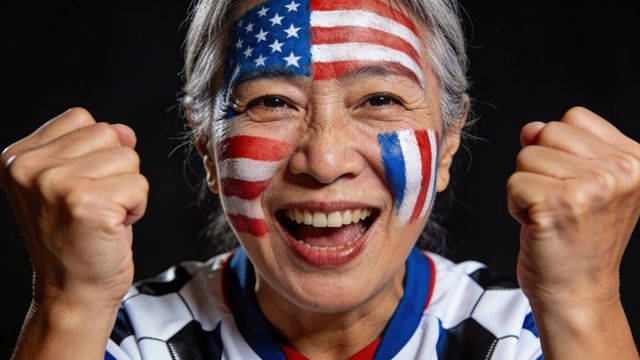 Energetic senior asian fan cheering with painted american flag face and raised fists