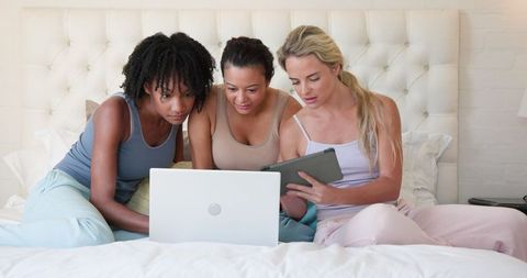 Diverse friends relaxing on bed using laptop and tablet