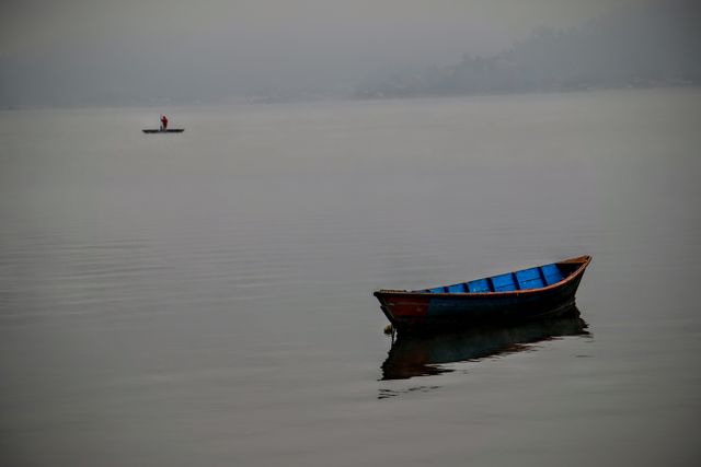Misty lake with wooden boat floating in peaceful water during early morning. Suitable for travel brochures, nature conservation campaigns, desktop backgrounds, relaxation apps, and artistic wall decor focused on tranquility and serene scenic beauty.