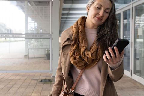 Mature woman checking smartphone at urban transit shelter wearing coat scarf crossbody bag