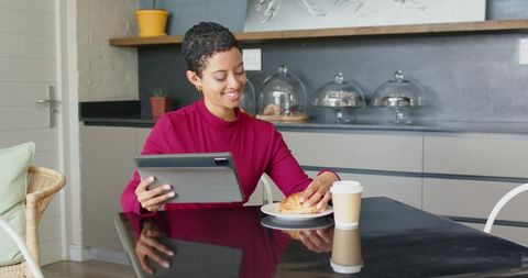 Smiling woman using tablet while enjoying croissant and coffee in modern kitchen