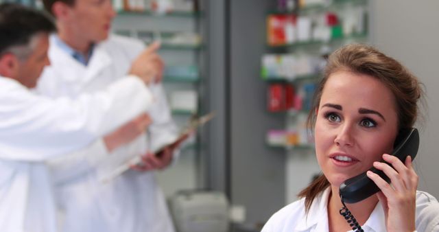 A young Caucasian woman is on the phone, a pharmacy technician, with two male pharmacists discussing in the background. Her attentive expression and the busy pharmacy setting suggest a focus on customer care and healthcare service.
