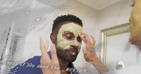Black man receiving facial mask from partner at home mirror, skincare self-care moment