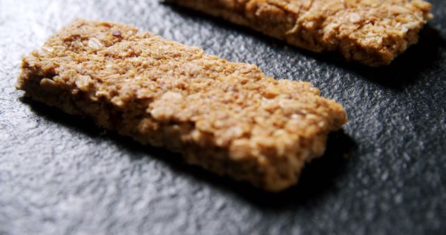 Two oatmeal bars are placed on a dark textured surface, offering a healthy snack option. Their rough texture and golden-brown color suggest a baked, wholesome treat rich in fiber.