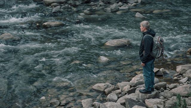 Hiker standing on rocky riverbank gazing at fast-moving mountain stream with backpack