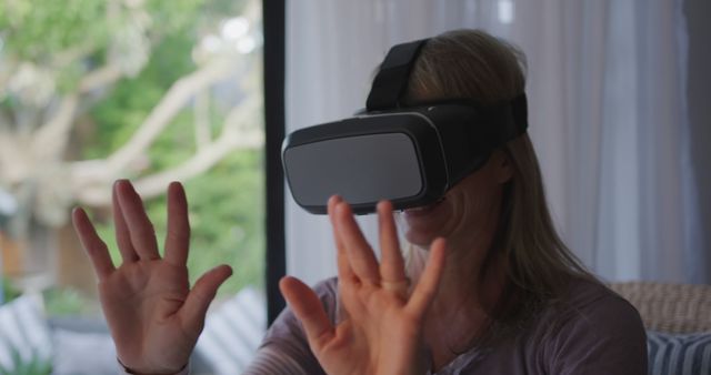 Woman using VR headset with her hands up, experiencing immersive technology indoors. Could be used to illustrate the future of digital interaction, modern relaxation, or advancements in virtual reality.