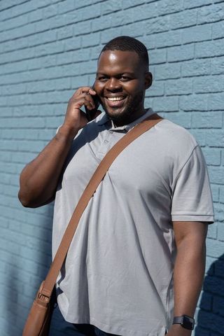 Smiling african american man talking on smartphone by blue brick wall with messenger bag