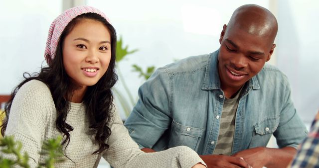 This stock photo shows two young adults, one wearing a pink hat, studying at home, contributing to a friendly and collaborative atmosphere. This image can be used for advertising educational services, promoting teamwork, online learning platforms, or highlighting diverse friendships.