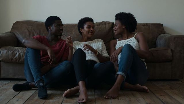 Three adults sitting on wooden floor chatting beside brown sofa, relaxed home conversation