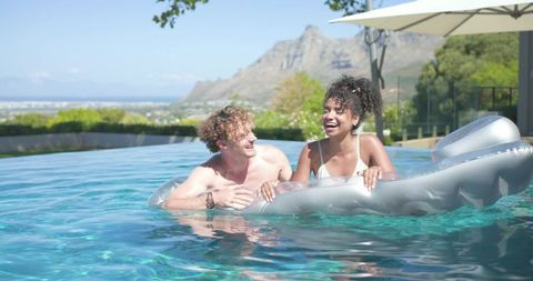 Laughing mixed-race woman and white man enjoying luxury infinity pool on sunlit coastal terrace