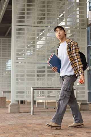 Asian student walking on modern urban campus holding notebooks backpack and apple