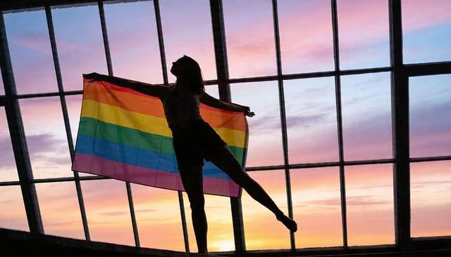 Silhouetted woman balancing on ledge holding rainbow flag in industrial loft soft sunset
