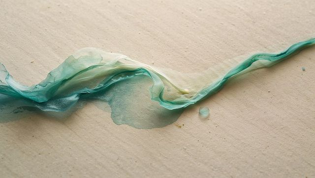 Flowing teal chiffon ribbon forming sinuous wave on textured beige paper with paint bleed