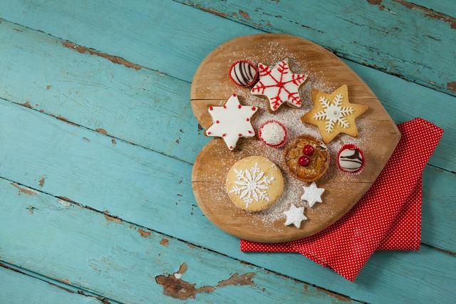 Christmas-themed gingerbread cookies decorated with icing placed on a heart-shaped wooden cutting board. Set on a rustic, blue wooden table with a red polka-dot cloth. Ideal for holiday greeting cards, seasonal advertising, baking blogs, and festive social media posts.