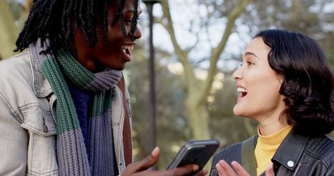 Diverse friends laughing and sharing smartphone while chatting in urban park with knit scarves
