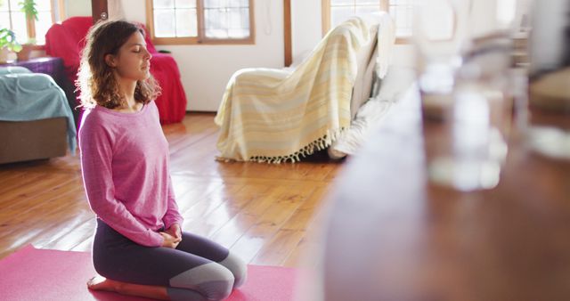 Relaxed biracial woman practicing yoga, kneeling on mat with eyes closed in sunny cottage bedroom. simple healthy living in off the grid rural home.