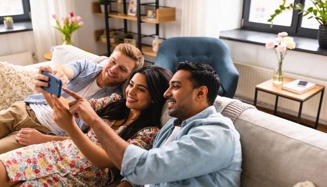 Three friends taking selfie on cozy sunlit living room sofa, laughing and leaning together