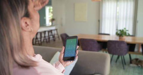Mature woman relaxing on sofa using smartphone with smart home app in open-plan lounge