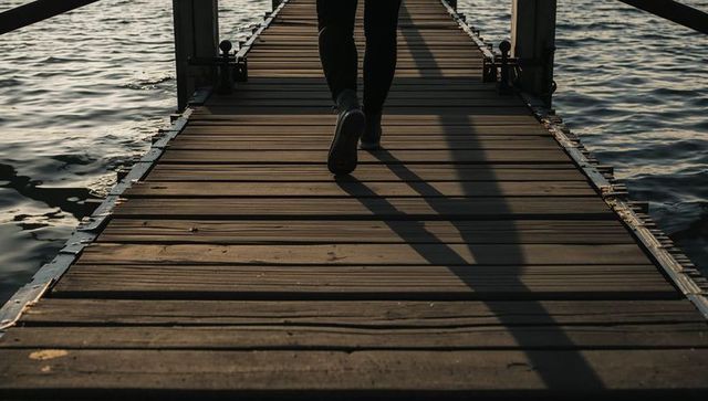 Walking on weathered dock at sunrise casting long shadow over calm water and wooden planks