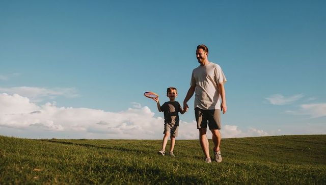 Father walking with son holding hands while carrying colorful frisbee on grassy hill