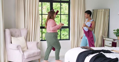 Diverse friends trying on clothes and chatting in bright cozy bedroom interior