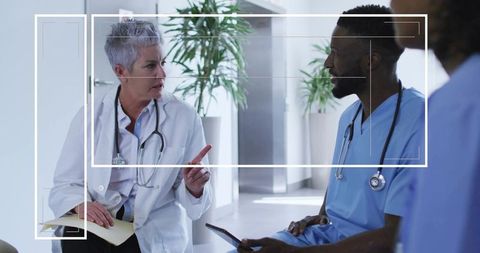 Senior physician consulting with medical team in hospital corridor using tablet and notes