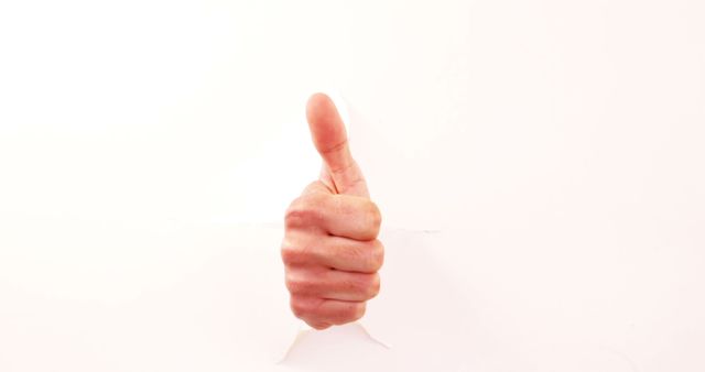 A Caucasian hand is shown giving a thumbs-up through a torn white paper, with copy space. This gesture typically signifies approval, agreement, or a job well done.