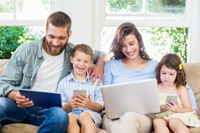 Family sitting on a sofa in the living room, each using a different digital device such as a laptop, tablet, and smartphone. The parents and children are smiling and appear to be enjoying their time together. This image can be used for promoting family-friendly technology, digital lifestyle, or home comfort products.