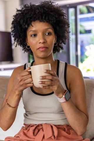African american woman sipping coffee on sofa, wearing smartwatch, serene morning at home