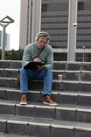 Middle-aged man sketching on tablet wearing headphones while sitting on urban concrete steps