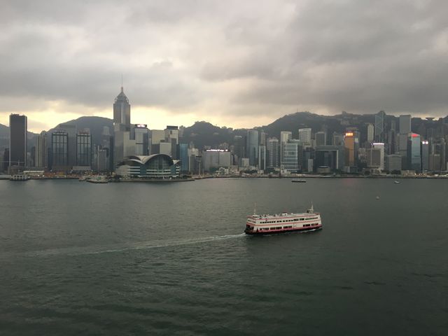 Captivating view of Hong Kong's iconic skyline with a ferry navigating Victoria Harbour against a cloudy sky. Ideal for travel promotions, city-related content, visual storytelling, transportation services, and urban landscape articles.