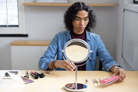 Nonbinary person arranging makeup at vanity mirror in sunlit workspace, beauty routine