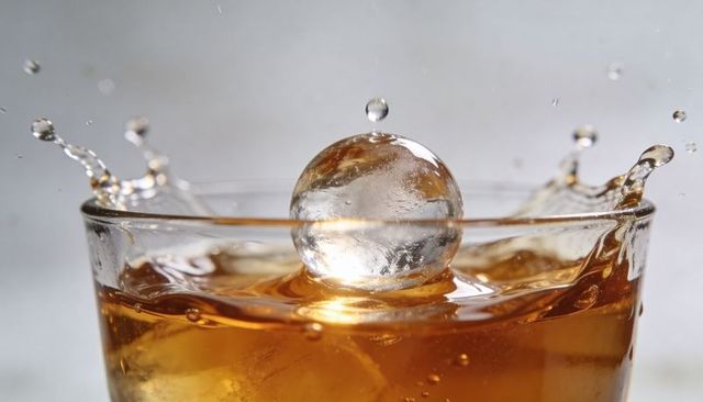 Amber splash with spherical ice cube in tumbler glass, macro crown splash motion