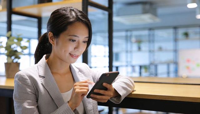 Asian woman checking phone while leaning on wooden counter in coworking space