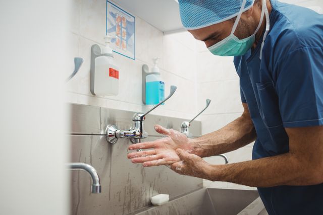 Male surgeon in blue scrubs and surgical mask washing hands in hospital. Emphasizes hygiene, cleanliness, and infection control in medical settings. Useful for healthcare, medical training, and hygiene awareness materials.