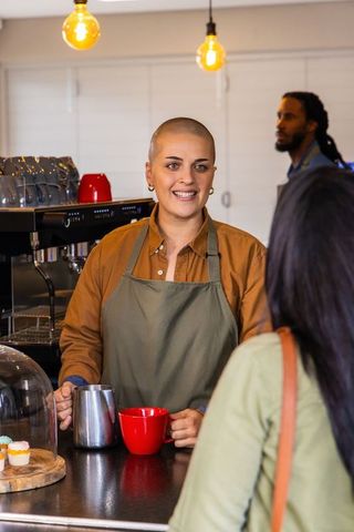 Barista serving coffee and smiling at counter with cupcakes and warm hanging lights