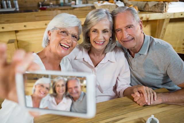 Three senior friends are smiling and taking a selfie with a mobile phone in a cozy cafe. They are sitting at a wooden table, enjoying each other's company. This image can be used for promoting senior lifestyle, social activities, friendship, and technology use among the elderly.
