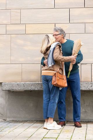 Senior couple kissing and embracing on city sidewalk, man holding bouquet, autumn fashion