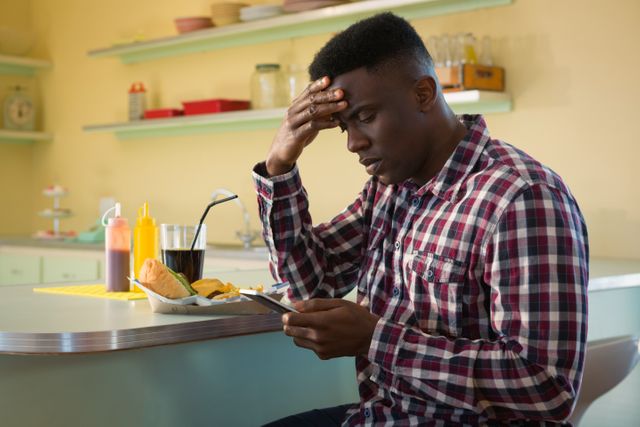 Man sitting at restaurant table, looking at mobile phone with worried expression. Casual setting with food and drink on table. Useful for themes of stress, technology, dining out, and modern lifestyle.
