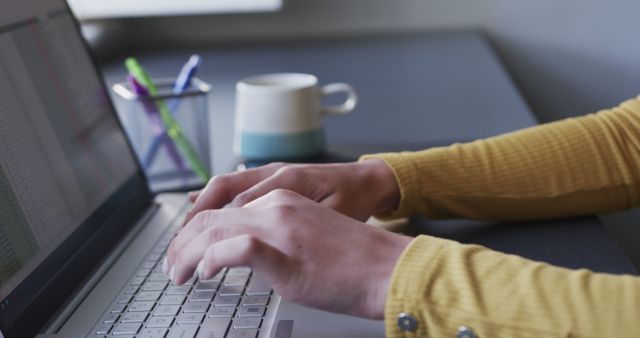 Person typing on laptop at desk with coffee mug and pen holder in background. Suitable for illustrating work environments, technology usage, productivity, remote working, academic or professional writing, and modern office settings.