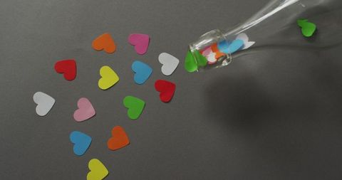 Colorful paper hearts scattered from glass bottle on gray surface