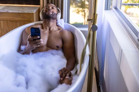 Mature african man relaxing in bubble bath listening to music with headphones and smartphone