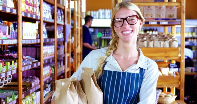 Young woman working in an organic grocery store, holding paper bags with a smile. Useful for depicting friendly customer service, small business promotion, healthy food shopping, and retail environments focused on organic and natural products. Shows the warmth and approachability of store staff in a well-stocked grocery aisle filled with natural and organic items.