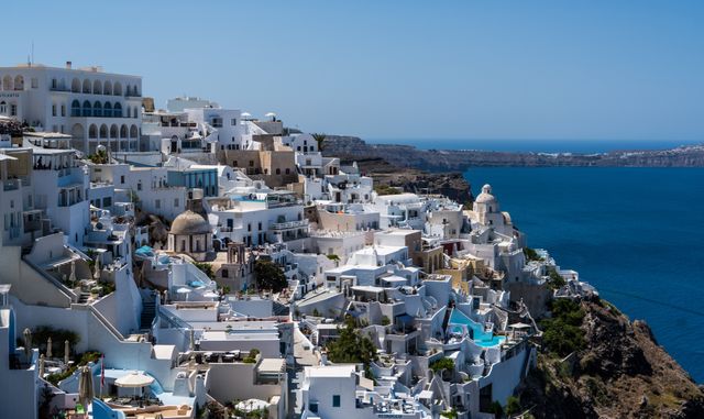 Picturesque view of whitewashed buildings clustered on cliffs overlooking the Aegean Sea under a clear blue sky. Ideal for promoting tourism, highlighting architectural beauty, or illustrating coastal and island life. Perfect for travel brochures, Mediterranean vacation ads, and lifestyle blogs.