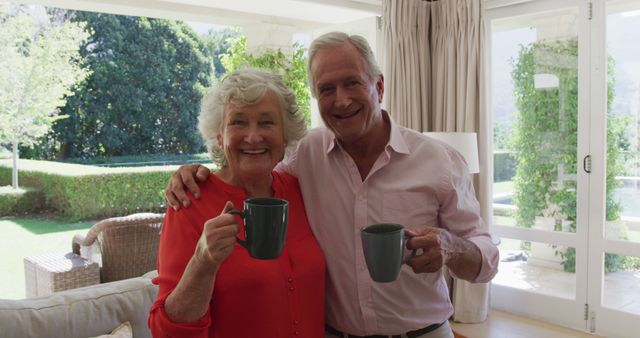 Portrait of happy caucasian senior couple embracing in sunny living room, smiling and holding cups. at home in isolation during quarantine lockdown.