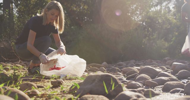 Caucasian female volunteer cleaning up a river on a sunny day in the countryside, picking up rubbish. Ecology and social responsibility in a rural environment.