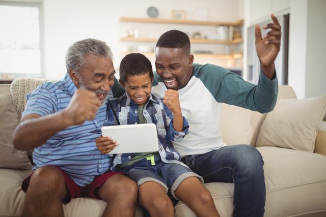 This image shows a joyful moment of a multi-generation family, including a grandfather, father, and son, using a digital tablet in a living room. They are visibly excited and celebrating, suggesting they might be watching a game or achieving something on the tablet. This image can be used for promoting family bonding, technology use in homes, or advertisements related to digital devices and family entertainment.
